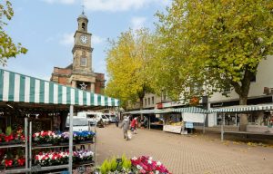Newcastle under lyme market - multiple stalls selling flowers and baked goods. The guildhall is in the background and the trees have an Autumnal look.