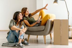 Two women sitting in a bright, unfurnished living room surrounded by moving boxes, discussing plans while one gestures ahead, suggesting a home move or relocation.