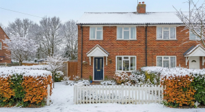 A red-brick semi-detached house with a white picket fence and front garden covered in snow, with bare trees and a frosty winter setting.