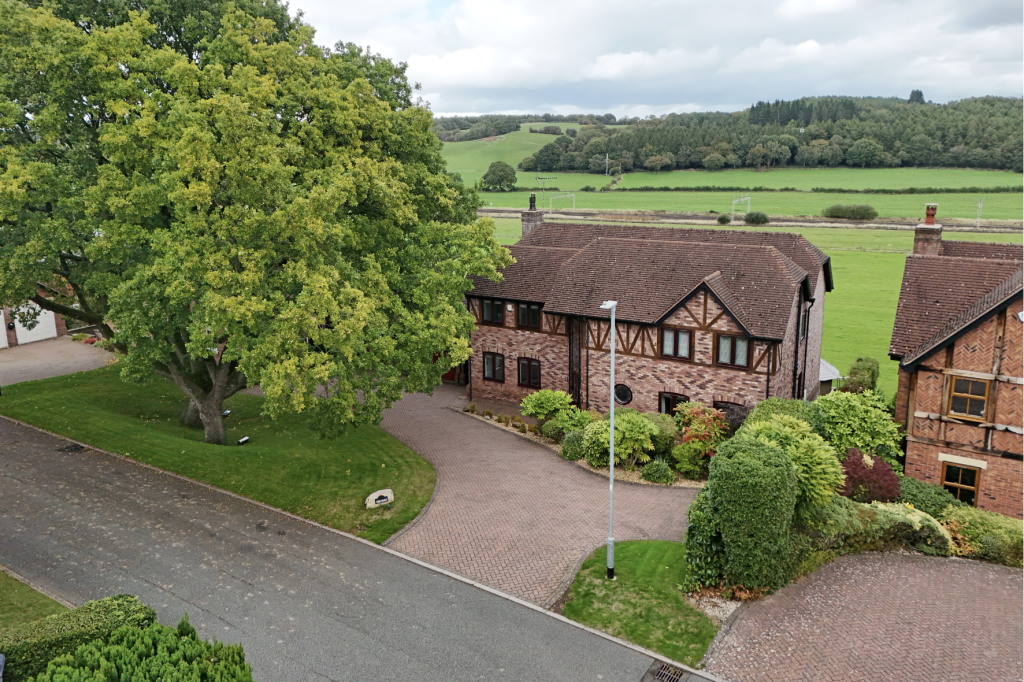 Detached brick house with driveway and mature tree, set against open countryside views in a quiet residential setting.